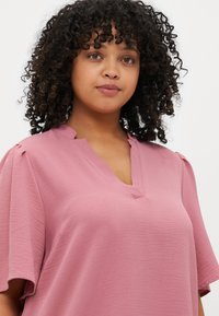 Pink short-sleeve blouse with a V-neckline and subtle texture. Features soft fabric, relaxed fit, and pleated detail on shoulders.
