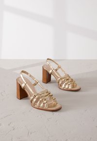 Gold strappy heeled sandals with a chunky wooden heel, featuring a woven upper design and adjustable ankle straps. Smooth metallic finish.