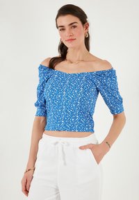 Blå blomsterprint crop top med off-the-shoulder design, korte puffærmer og en tætsiddende talje. Sammen med hvide bukser med snoretræk.
