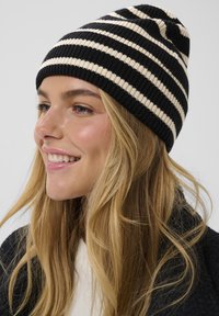 Berretto a maglia a strisce nere e crema con texture a costine; design aderente; indossato leggermente slacciato da una persona con capelli lunghi e ondulati.