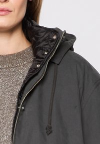 Black hooded jacket with a zip closure, featuring a quilted inside lining and drawstring accents. Worn over a textured, sparkly sweater.
