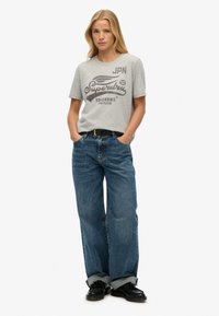 T-shirt en coton gris avec un texte graphique noir, associé à un jean en denim bleu à jambes larges. La tenue est complétée par des chaussures noires et une ceinture.