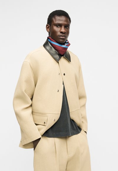 Homme portant une veste beige et un pantalon assorti avec un col noir, une chemise noire et un foulard rayé rouge et bleu autour du cou, posant les mains dans les poches.