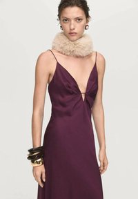 Vestito di raso prugna con scollatura a V profonda e dettaglio a nodo sul petto. Presenta spalline sottili ed è abbinato a un colletto di pelliccia leggera. Bracciali neri e dorati.