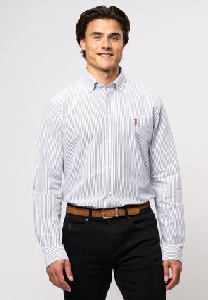 Hemd mit Knopfleiste in navy und weißen vertikalen Streifen, langen Ärmeln und einem kleinen gestickten Emblem auf der Brust. Getragen mit schwarzen Jeans und einem braunen Gürtel.
