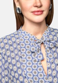 Blaue Bluse mit floralen Mustern, einer Krawattenhalsdetail und kontrastierenden Designs. Große silberne Ohrstecker runden den Look ab.