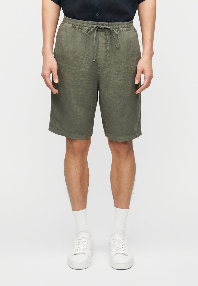 Homme portant un short en lin vert olive à cordon, des chaussettes blanches montantes et des baskets blanches, debout devant un fond uni.