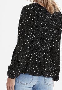 Schwarze Bluse mit Punkten, die einen gerafften Rücken, lange Puffärmel und einen ausgestellten Saum aufweist. Der Stoff wirkt leicht und hat eine weiche Textur.