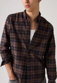 Καφέ και ναυτικό καρό φανελένιο πουκάμισο με κουμπιά μπροστά, μακριά μανίκια γυρισμένα στις μανσέτες και απαλό υφασμάτινο, φορεμένο πάνω από ένα λευκό τοπάκι.