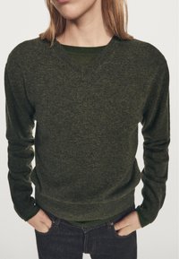 Grøn v-hals sweater lavet af et tekstureret strikket stof, med lange ærmer og en afslappet pasform, kombineret med en mørkere grøn langærmet skjorte under.