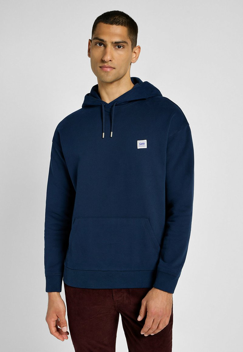 Sudadera azul marino de algodón, con un bolsillo delantero, capucha ajustable con cordón, puños de canalé y un pequeño parche con logo en el pecho.