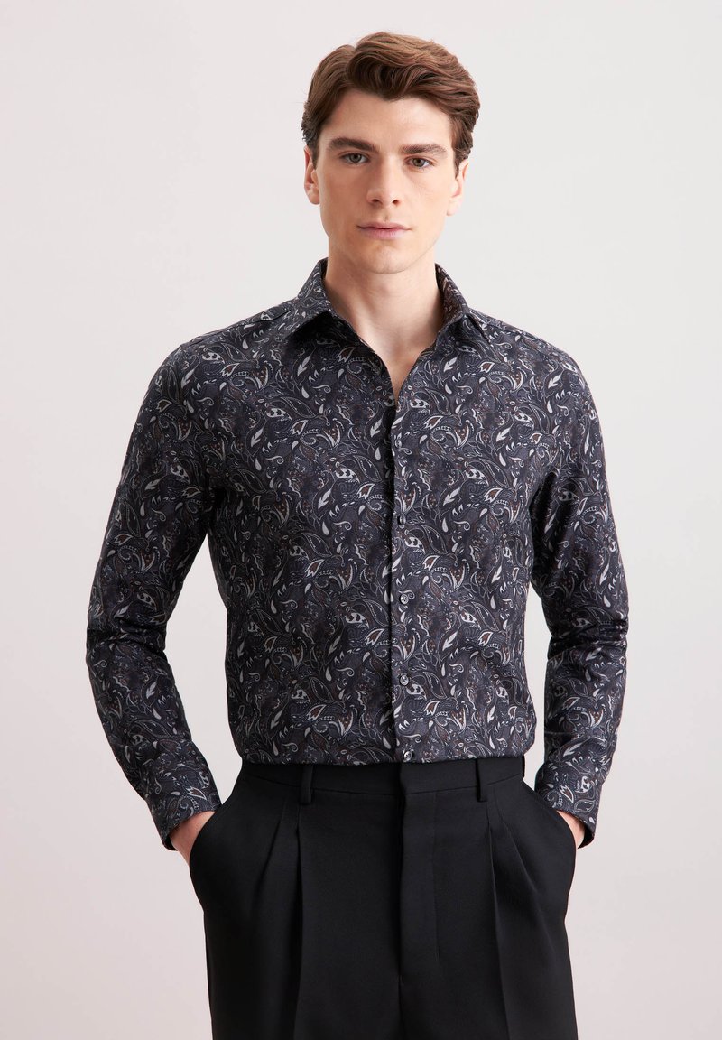 Camicia nera a maniche lunghe con un motivo paisley in grigio e bianco. Presenta un colletto classico e una chiusura con bottoni. Indossata con pantaloni neri su misura.