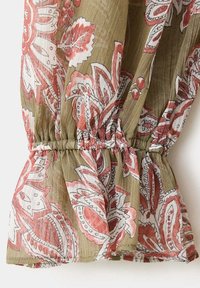 Gros plan d'un tissu avec un fond vert olive et de grands motifs floraux et cachemire rouges et blancs, avec un détail élastique froncé au centre.