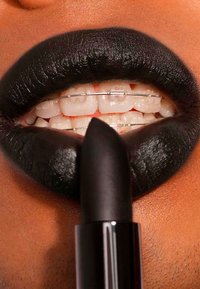 Svart leppestift påførte lepper med glossy tekstur, som viser hvite tenner og tannregulering. Leppestift-røret er svart med en spiss tupp.