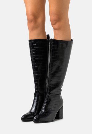 Jambes portant des bottes en cuir noir à texture crocodile montant jusqu'aux genoux, avec des talons carrés, se tenant sur un fond gris clair uni.