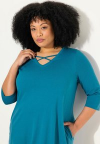 Abito teal a tre quarti con scollo a V e bretelle incrociate sul collo. Tessuto morbido con una vestibilità rilassata e tasche laterali.