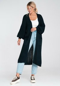 Długi, ciemnozielony, dziany kardigan z szerokimi rękawami, z kieszeniami z przodu, otwarty z przodu, noszony na białej bluzce bez rękawów i jasnoniebieskich dżinsach.