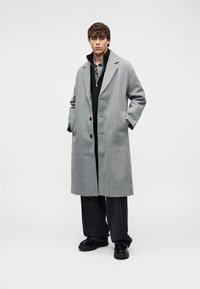 Γκρι μάλλινο παλτό με μεγάλους γιακάδες, δύο κουμπιά και πλαϊνές τσέπες, φορεμένο πάνω από μαύρο φερμουάρ και ριγέ πουκάμισο, συνδυασμένο με σκούρο παντελόνι και μαύρα παπούτσια.