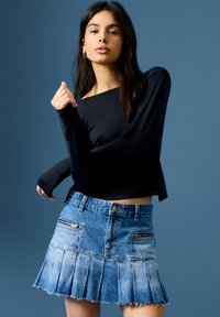 Črn dolg rokav vrh v kombinaciji s svetlo modrim denim plisiranim krilom, ki ima dva žepa na zzip in raztrgan rob. Modra ozadje.