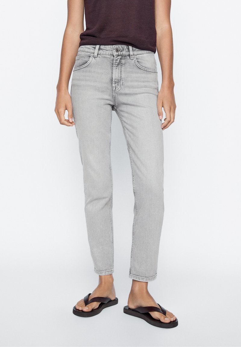 Jean en denim gris clair avec une coupe slim, taille moyenne, design à cinq poches et coutures contrastées. Associé à des tongs noires.