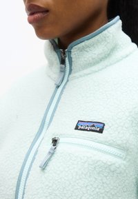 Κλείσιμο λήψης ενός ατόμου που φοράει ανοιχτό μπλε φλις μπουφάν Patagonia με φερμουάρ στο τσεπάκι του στήθους και ψηλό, φερμουάρ κλειστό κολάρο.