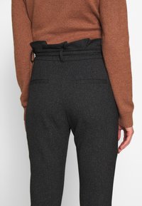 Black tailored trousers with a textured finish, featuring a high ruffled waistband and two back pockets, paired with a brown sweater.