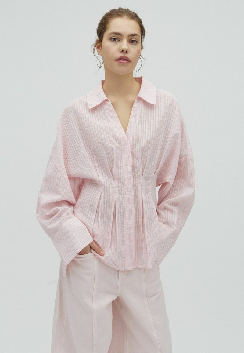 Jeune femme portant un chemisier ample rayé rose clair avec des manches larges et un pantalon rose clair, debout avec les mains dans les poches.