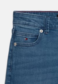 Modré džínové kalhoty se přední kapsou s malou logovou nálepkou, stříbrnými doplňky a kontrastním červeným prošíváním kolem pasu.