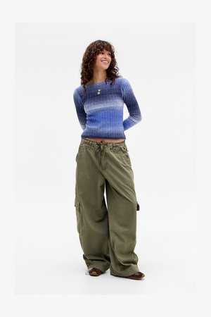 Donna con capelli ricci che indossa un maglione a righe blu ombré, pantaloni cargo larghi verde oliva e scarpe marroni, in piedi con le mani dietro la schiena.