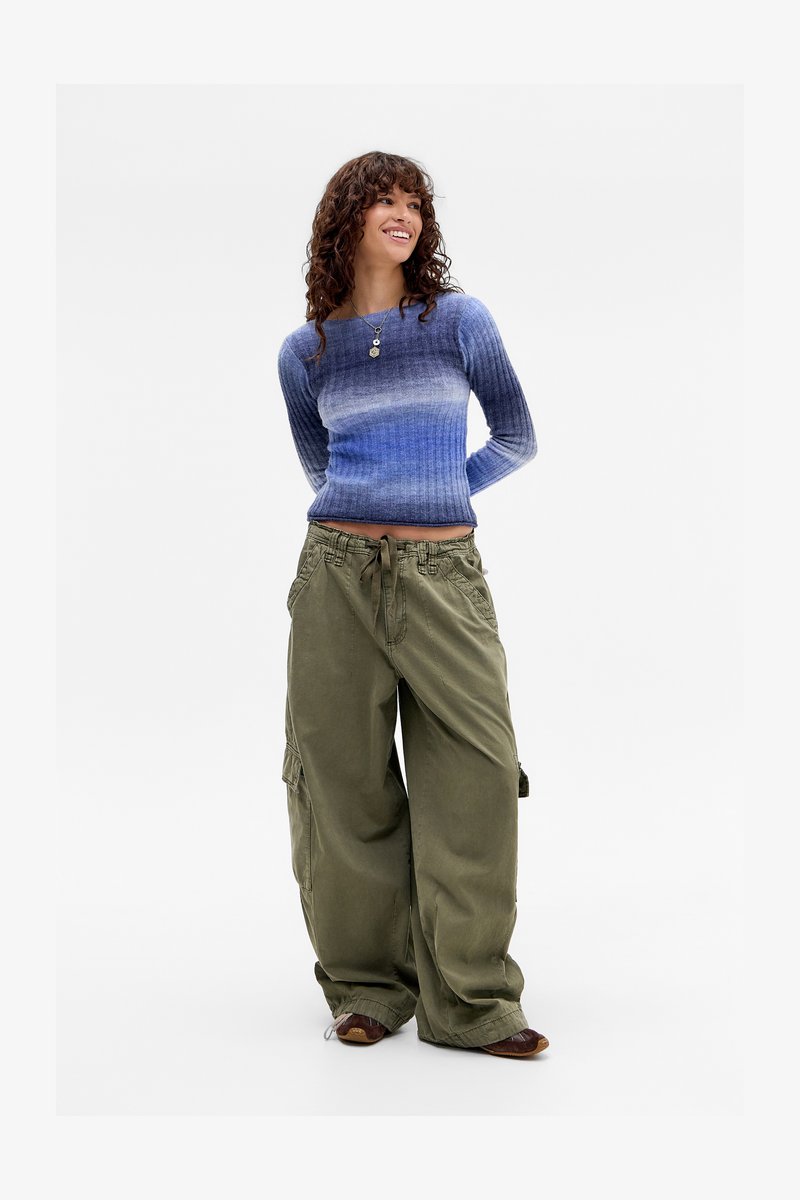 Frau mit lockigem Haar, die einen blau-ombre gerippten Pullover, olivgrüne, weite Cargo-Hosen und braune Schuhe trägt und mit den Händen hinter dem Rücken steht.