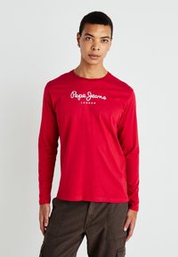 Fiatal férfi, aki piros hosszú ujjú Pepe Jeans London inget és sötét munkanadrágot visel, egy sima világos háttér előtt áll.