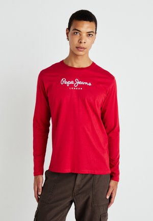 Joven vestido con una camisa de manga larga roja de Pepe Jeans London y pantalones cargo oscuros, de pie contra un fondo claro y liso.