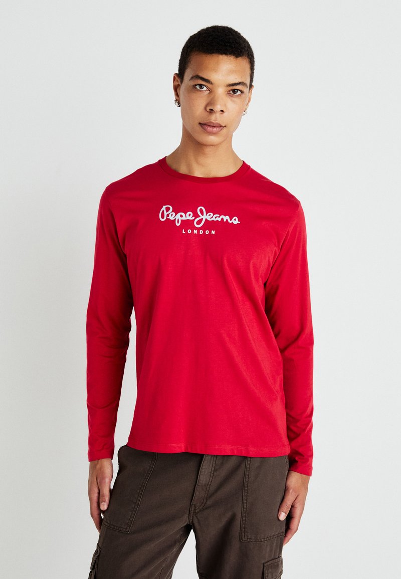 Fiatal férfi, aki piros hosszú ujjú Pepe Jeans London inget és sötét munkanadrágot visel, egy sima világos háttér előtt áll.