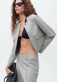 Veste cropped gris clair en tissu doux avec des détails de boutons en or, associée à un haut de bikini noir et un sac à main tissé noir.