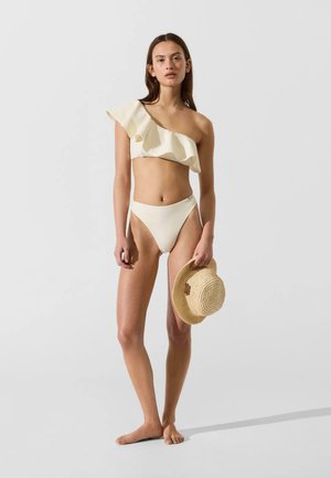 Donna in piedi a piedi scalzi, indossa un bikini crema monospalla con volant, tiene un cappello di paglia sullo sfondo chiaro e uniforme.