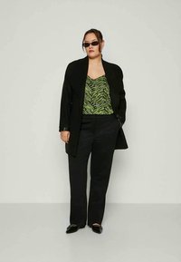Mulher a usar óculos de sol pretos, casaco preto, blusa verde com padrão, calças pretas e sapatos pretos, de pé contra um fundo claro e liso.