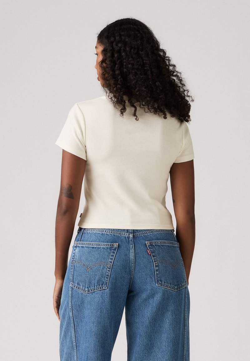 T-shirt crème à manches courtes avec un design ajusté, associé à un jean en denim bleu taille haute avec des poches arrière et un onglet logo rouge.