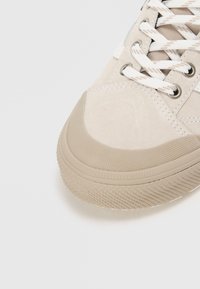 Bež nubukist sneaker ümmarguse toe, tekstureeritud kummitald, valged paelad ja väikesed heleda pruuni tooni õmblused.