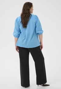 Blouse bleu clair avec motif texturé, manches bouffantes, et pantalon large noir. La blouse présente un ourlet arrondi et un détail d'empiècement au dos.