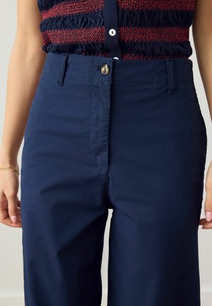 Frau trägt hoch taillierte marineblaue Hose mit Knopf und Reißverschluss, kombiniert mit einem ärmellosen marineblauen und roten Stricktop.