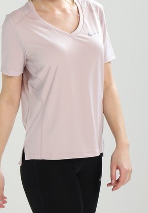 Frau trägt hellrosa Nike T-Shirt mit V-Ausschnitt und kurzen Ärmeln sowie schwarze Leggings vor einfarbigem Hintergrund.