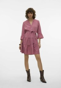 Rozā rakstainā kleita ar trim ceturtdaļām garām piedurknēm, V-veida kakla izgriezumu, izsistām vidukli un daudzslāņu svārkiem, kopā ar brūnām augstpapēžu zābakiem.