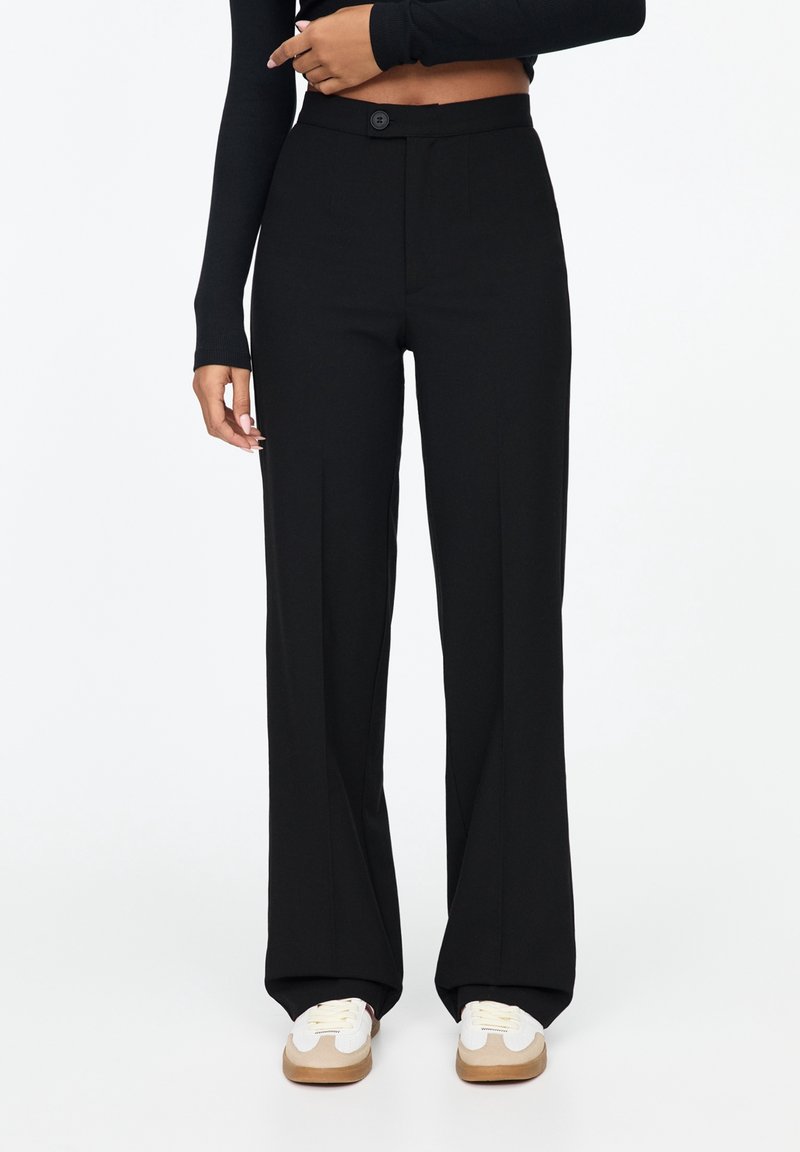 Femme portant un pantalon noir taille haute à jambes larges, associé à des baskets blanches et un top court noir à manches longues, debout sur fond blanc.