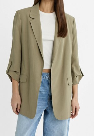Femme portant un blazer oversize vert olive clair avec les manches retroussées, un top court blanc côtelé et un jean bleu clair sur fond uni.
