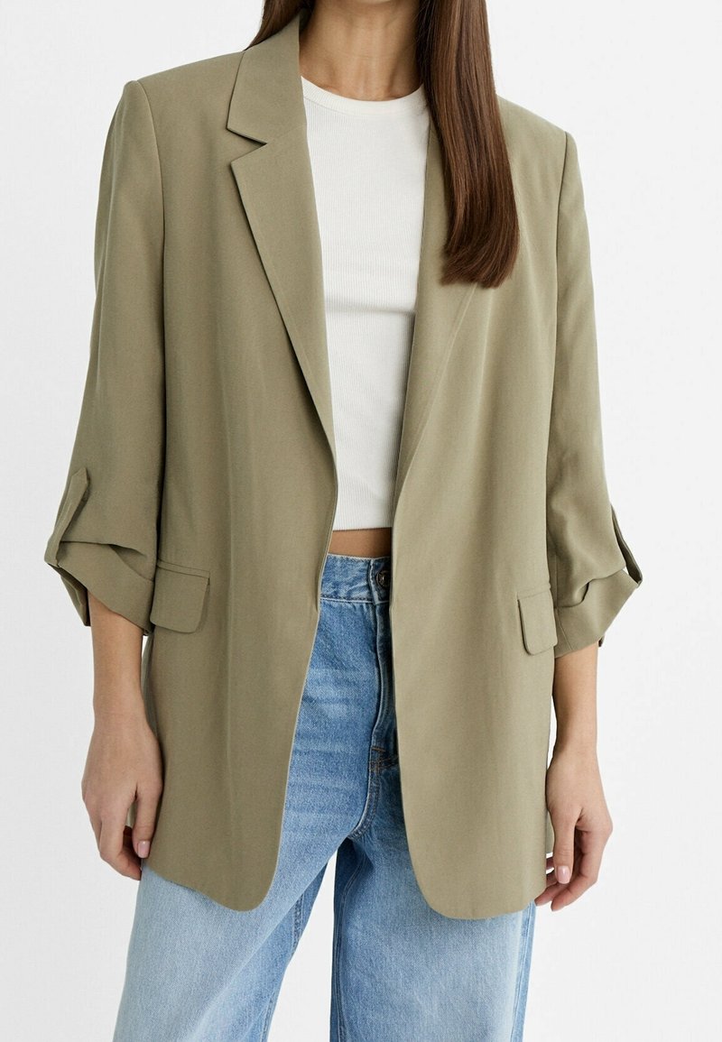 Femme portant un blazer oversize vert olive clair avec les manches retroussées, un top court blanc côtelé et un jean bleu clair sur fond uni.