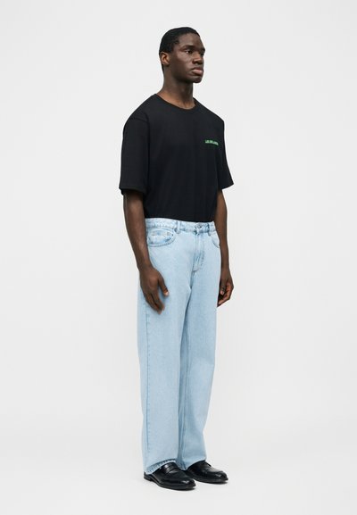 Młody mężczyzna stojący, ubrany w czarną koszulkę z zielonym napisem, jasnoniebieskie dżinsy o prostych nogawkach oraz czarne skórzane buty, na tle jednolitego jasnego tła.