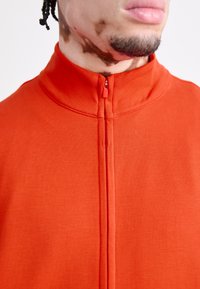 Casaco vermelho com fecho zip e colarinho alto, feito de um tecido liso, com um fecho central à frente.