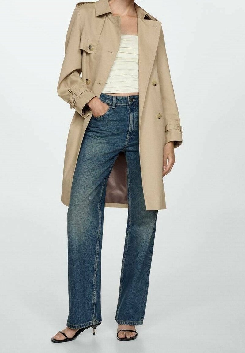 Manteau beige à double boutonnage, associé à un haut blanc ajusté et un jean bleu à jambe large. Des sandales noires viennent compléter le look.