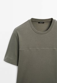 T-shirt verde oliva con scollatura rotonda e maniche corte. Presenta una sottile cucitura orizzontale sul petto e una texture di cotone liscia.