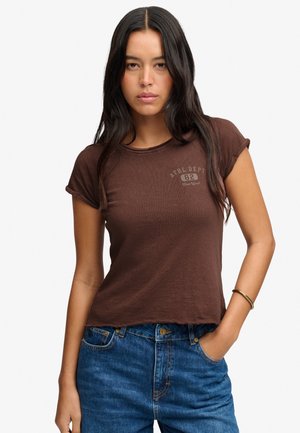Mujer con cabello largo y oscuro que lleva una camiseta marrón de manga corta y vaqueros azules, con una mano en el bolsillo y una pulsera en la muñeca.